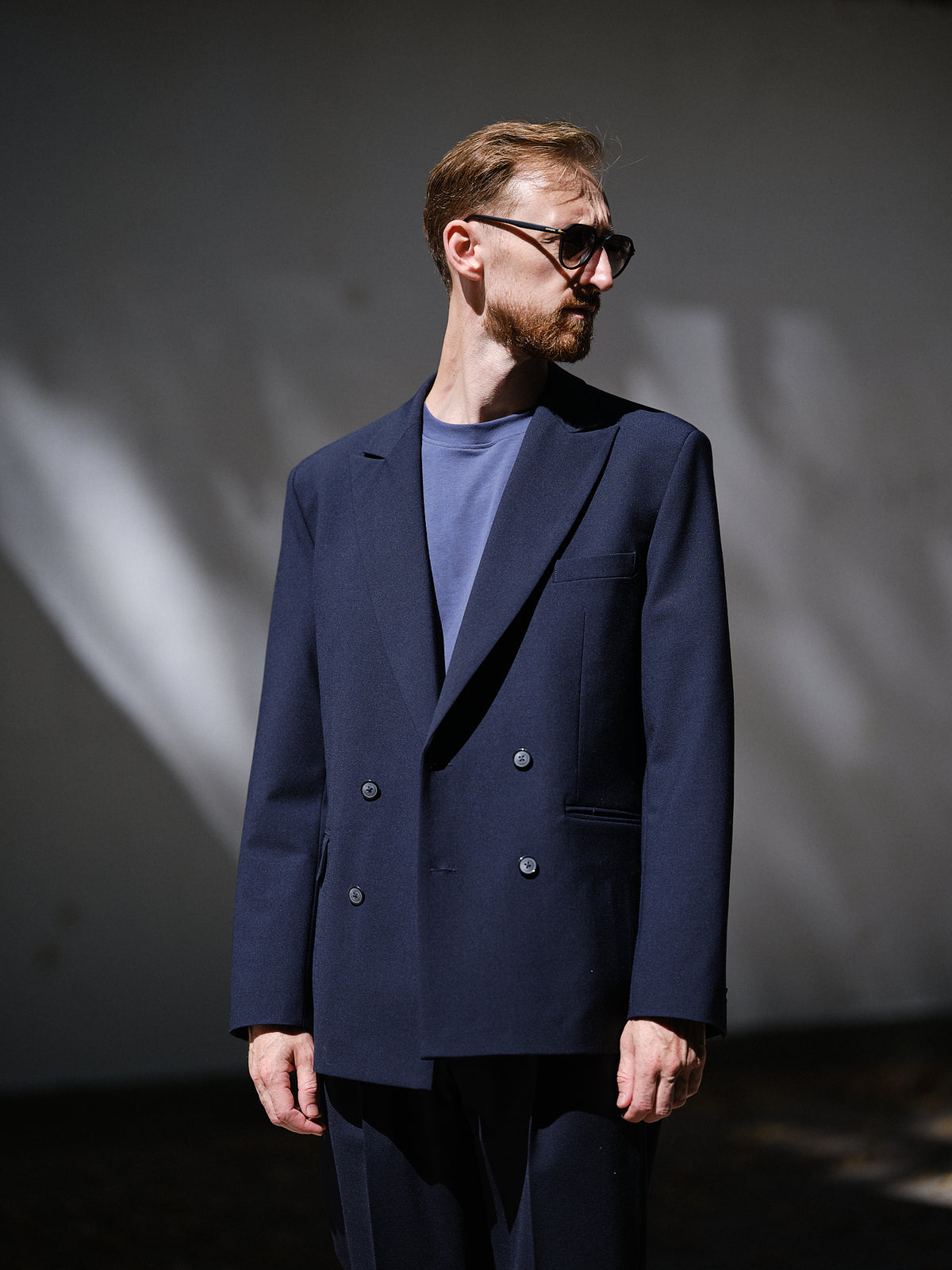 A man wearing a navy blue double-breasted blazer over a slate blue crewneck t-shirt, styled with dark sunglasses, standing against a dark gray wall with dramatic light shadows.