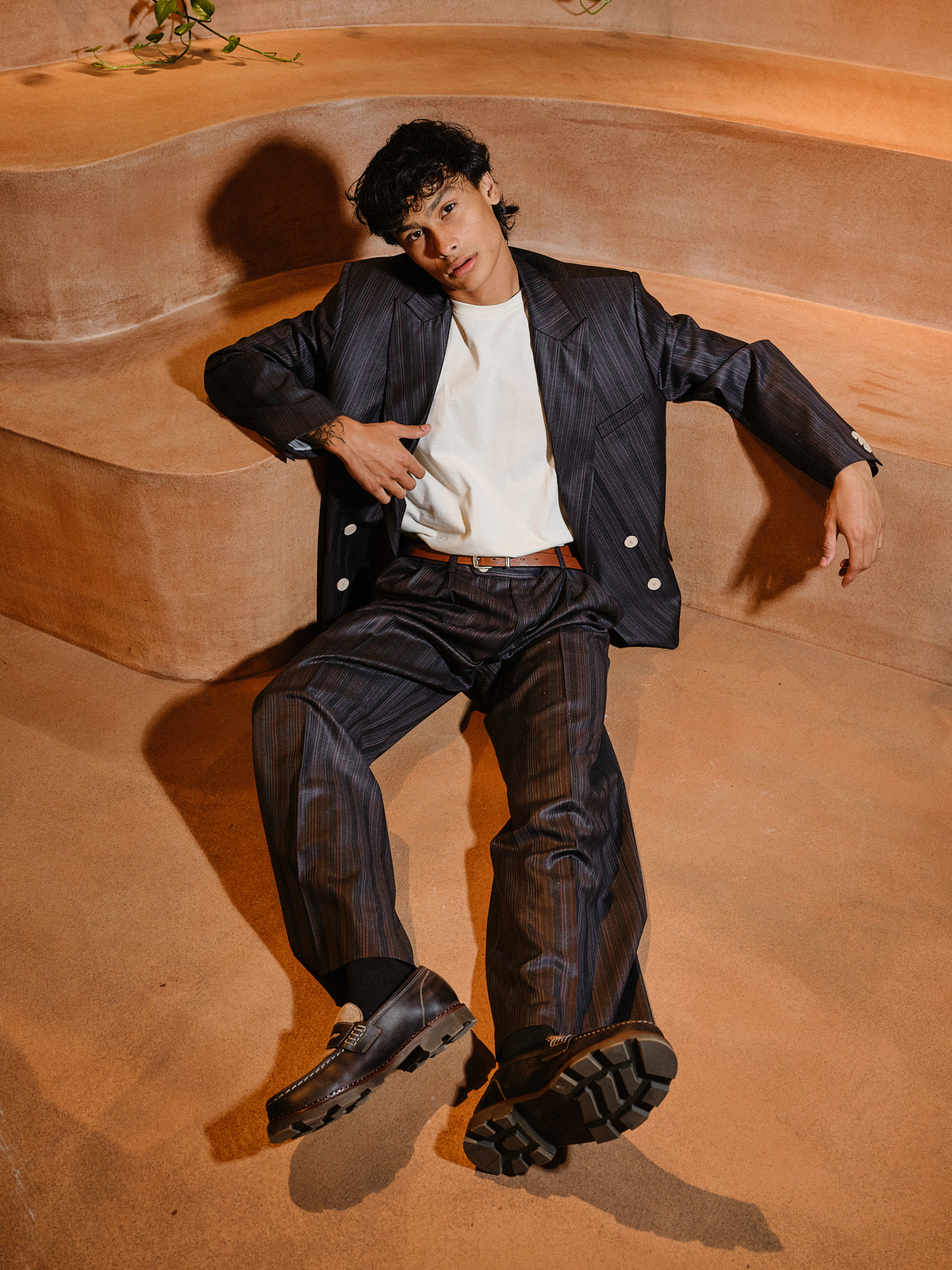 A man wearing a dark navy pinstripe blazer and matching trousers over a white t-shirt, sitting on terracotta-colored concrete steps.